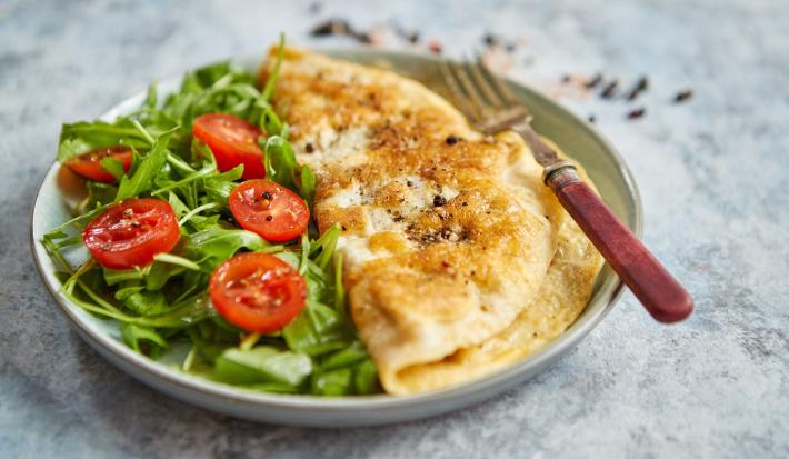 Foto da Receita de Omelete Baveuse. Observa-se um prato com a omelete dobrada no meio, servida com salada de rúcula e tomatinhos cerejas cortados ao meio.