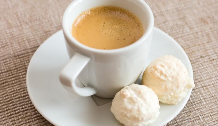 fotografia em tons de branco, cinza e marrom de uma bancada cinza. Contém um pratinho redondo branco com uma xicara de café branca com café dentro, ao lado dois bolinhos redondos.