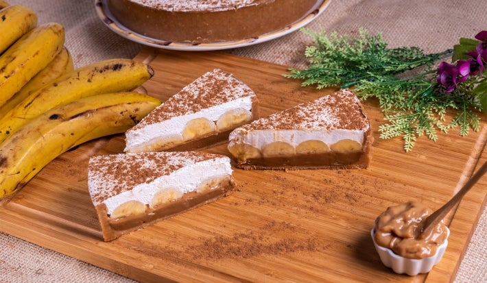 Em cima de uma mesa, fatias de banoffee com massa de biscoitos crocante, doce de leite, bananas em rodelas, decorado com chantilly cremoso, polvilhado canela em pó.