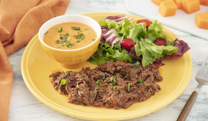 Foto da receita de Purê de Abóbora com Carne Desfiada. Observa-se um prato amarelo com a carne desfiada e a salada e, dentro de um potinho amarelo, o purê de abóbora finalizado com salsinha.
