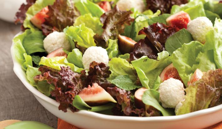 Uma bancada de madeira cinza vista de cima contém um pano laranja, um recipiente redondo e branco com salada dentro pedaços de figo e bolinhas de ricota. À frente duas colheres verdes para mexer a salada.