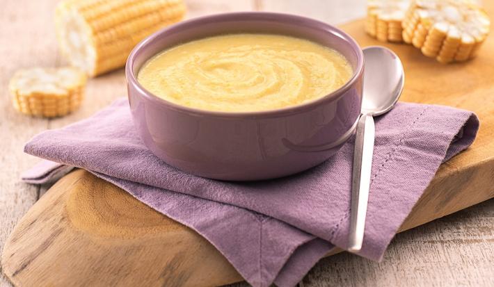 Foto em tons de amarelo da receita de caldinho de milho-verde servida em um bowl de porcelana roxo sobre um paninho roxo também com uma colher prateada ao lado. Tudo sobre uma tábua de madeira e ao fundo espigas de milho cortadas em rodelas