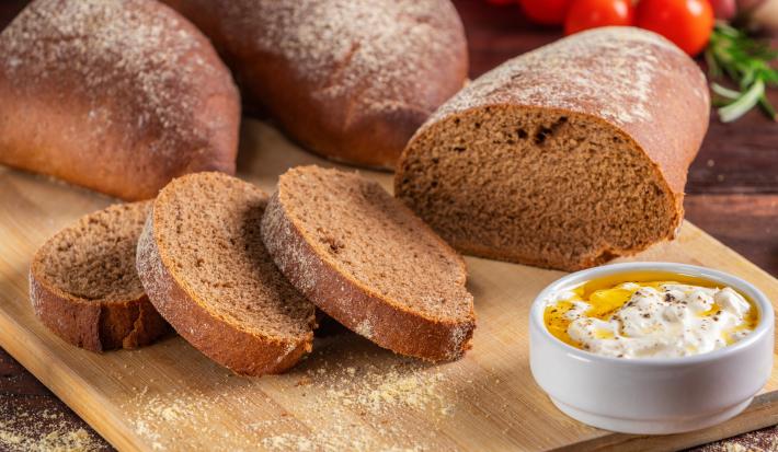 Fotografia de uma tábua média de madeira sobre uma mesa de madeira escura. Sobre a tábua tem uma receita de pão australiano com três fatias cortadas e sobrepostas ao lado de dois pedaços maiores do pão.