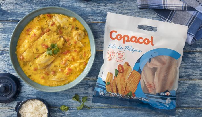 Foto da receita de Moqueca Fácil com Filé de Tilápia Copacol, vista de cima, servida em uma tigela azul clara, sobre uma bancada azul com alguns elementos de comida, um tecido e um pacote do produto Copacol
