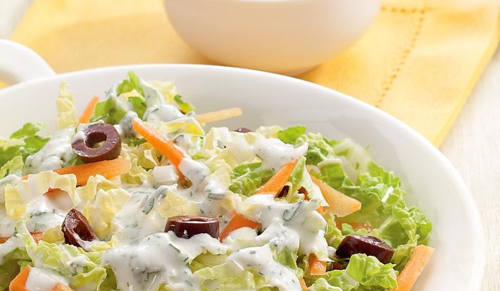Fotografia em tons de tons de branco, amarelo e verde, saladeira com folhas verdes, azeitona, cenoura e molho de iogurte sobre guardanapo amarelo, ao lado potinho com molho de iogurte e folhas de hortelã, tudo sobre bancada em tom pastel.