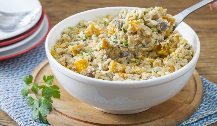 Fotografia em tons de branco, amarelo e marrom, com bowl branco ao centro sobre tábua de cozinha, ramo de salsa e um guardanapo azul, com arroz cremoso com legumes e carne picada, ao fundo pratos com garfos por cima, tudo sobre bancada de madeira marrom.