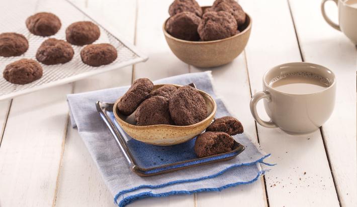 Fotografia em tons de azul em uma bancada de madeira clara, um paninho azul, um potinho bege com alguns cookies de cacau dentro dele. Ao fundo, mais potinhos com cookies e xícaras brancas com café para acompanhar.