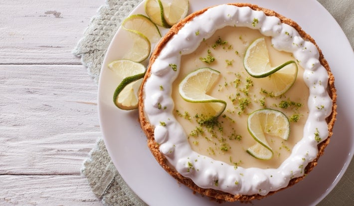 Fotografia em tons mais claros de uma torta inteira de limão com raspas e rodelas finas de limão. O chantilly está apenas na borda da torta, ao lado dela tem três fatias, que está sobre um prato grande e raso de vidro.