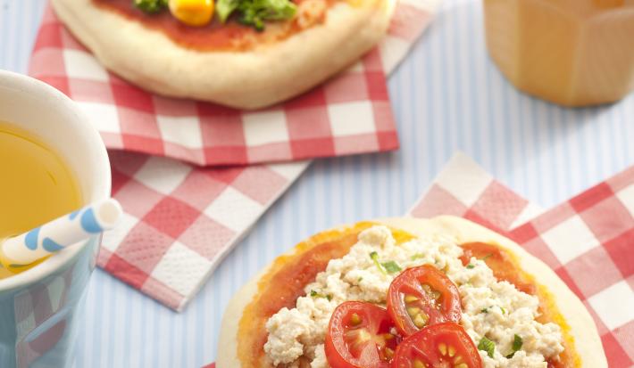 Fotografia em tons de vermelho em uma bancada cinza com guardanapos vermelhos em xadrez e uma mini pizza de legumes e outra mini pizza de tofu apoiadas sobre eles. Ao lado, um copo laranja com suco de laranja e um canudo branco com bolinha azul.