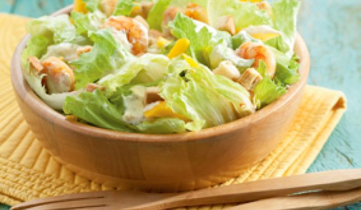 Fotografia em tons de azul, amarelo e marrom de uma bancada azul com um paninho amarelo, sobre ele um bowl de madeira com salada e uma colher e um garfo de madeira. Ao fundo pratos redondos brancos.