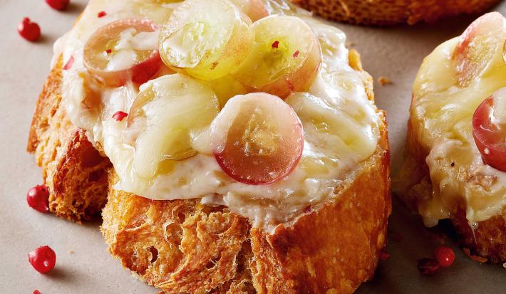 Fotografia em tons de amarelo em uma bancada de madeira escura com um prato branco raso e três bruschettas com queijo brie e uvas.