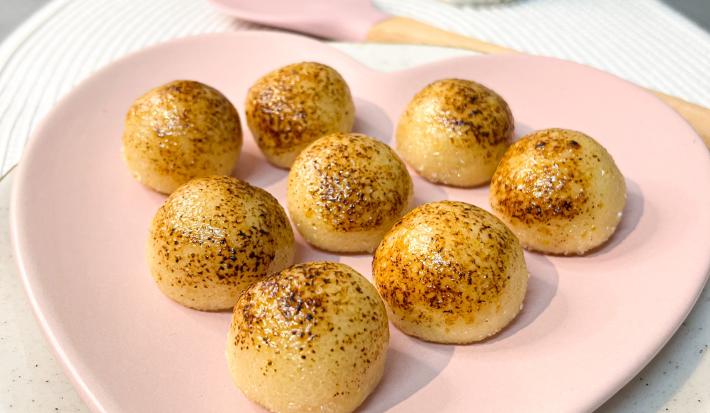 Foto da receita de brigadeiro brulle da andressa ventura, servido em um prato rosa em formato de coração, numa mesa onde há uma lata de leite moça, um pequeno vaso de flores