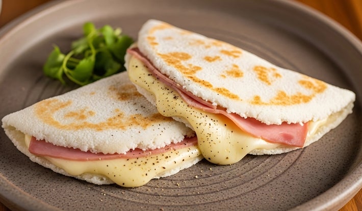 Fotografia de duas tapiocas pequenas recheadas com presunto, queijo e orégano, sobrepostas, em um prato raso e grande de tom marrom.