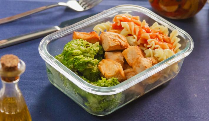 Fotografia mostra uma marmita em pote de vidro organizada com macarrão penne ao molho de tomate vibrante no canto superior direito, cubos de frango dourados ao meio, e brócolis verde intenso no espaço inferior.