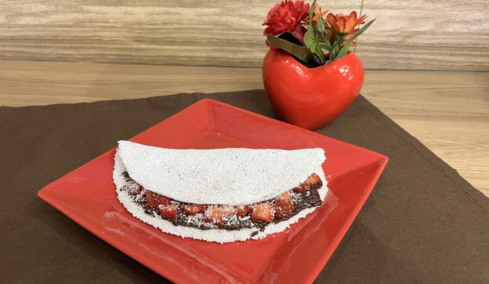 Fotografia em tons de vermelho e marrom com um prato vermelho ao centro. Em cima do prato existe uma receita de tapioca recheada de creme de amendoim e NESCAU Extra Cacau 60%