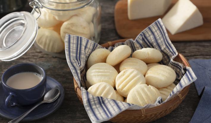 Fotografia em tons de azul em uma bancada de madeira escura, uma cesta de vime com um guardanapo de papel azul listrado com biscoitos de queijo dentro dele. Ao lado, uma xícara de café. Ao fundo, uma tábua de madeira com um pedaço de queijo.