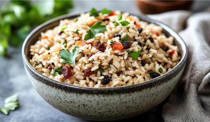 Em cima de uma mesa, tigela com arroz soltinho uvas-passas pretas e brancas, decorado com folhas de cheiro-verde.
