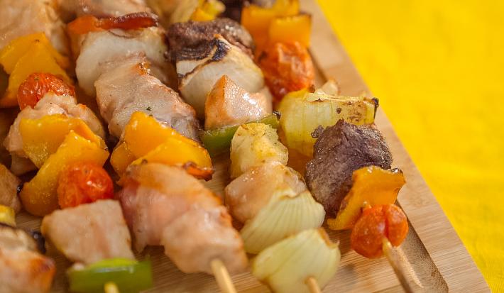 Imagem bem aproximada de uma tábua de madeira com cinco espetinhos sobre ela, alguns com frango, outros com carne e lombo. Eles também têm vegetais em tons verde, amarelo e vermelho.