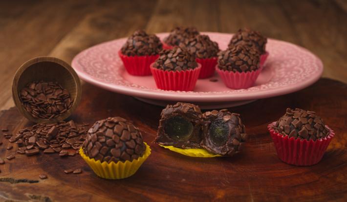 Fotografia em tons de amarelo e vermelho em uma bancada de madeira escura, um prato redondo rosa com brigadeiros em forminhas vermelhas em cima dele. Ao lado, brigadeiros em forminhas amarelas recheados com uva. Fotografia em tons de amarelo e vermelho em uma bancada de madeira escura, um prato redondo rosa com brigadeiros em forminhas vermelhas em cima dele. Ao lado, brigadeiros em forminhas amarelas recheados com uva.