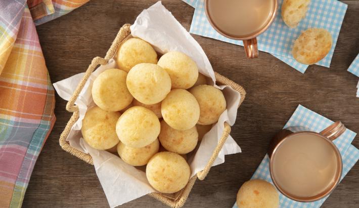 Imagem vista de cima de uma cesta com pães de queijo. Ao lado, 2 xícaras de café com leite e pães de queijo sobre um guardanapo.