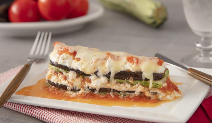 Foto da Receita de Lasanha de Ratatouille com Creme de Ricota. Observa-se um prato quadrado branco com uma fatia da lasanha. Atrás, de fundo, tomates e abobrinha decoram a foto.