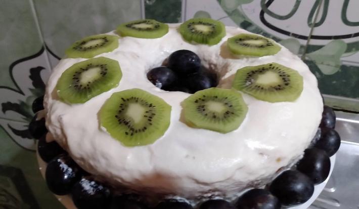 Foto da receita de bolo de cenoura confeitado servido com calda de chantilly, rodelas de kiwi cortadas por cima e uvas rosas para decorar em volta