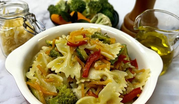 Foto em tons de amarelo da receita de farfalle com legumes servida em uma panela pequena de porcelana vermelha e branca. Ao fundo um vidro de azeite, um triturador de sal, uma porção de legumes e um pote de vidro com farfalle