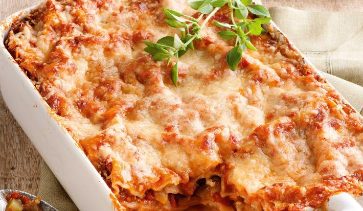 Fotografia em tons de branco e vermelho, ao centro refratário com lasanha de legumes gratinada com queijo, ao lado guardanapo bege com colher de servir, ao fundo prato branco com borda vermelha e porção de lasanha, tudo sobre bancada de madeira rústica.
