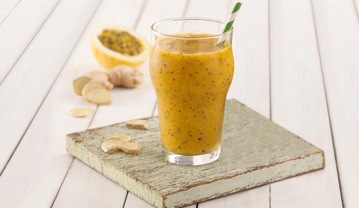 Fotografia em tons de amarelo em uma bancada de madeira branca, um paninho verde, uma tábua e madeira, um copo de vidro alto com o smoothie de manga com gengibre dentro dele. Ao fundo um pedaço de maracujá.