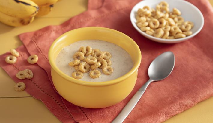 Fotografia em tons de amarelo em uma bancada de madeira amarela com um paninho laranja, um potinho amarelo com o creminho de banana e Cheerios dentro, uma colher e um potinho branco com os cereais Cheerios.