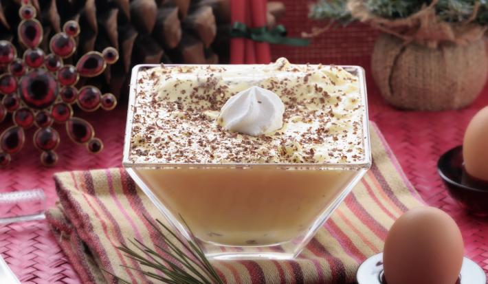 Fotografia de um flan de NESCAFÉ ao molho zabaione dentro de um recipiente quadrado de vidro fundo, com raspas de chocolate e chantilly por cima. A sobremesa está sobre um pano de mesa colorido.