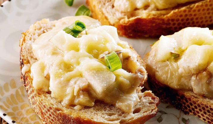 Fotografia em tons de creme, marrom e branco mostrando fatias de pão cobertas com um creme de queijo e cebolinha sobre bandeja com motivos florais, tudo sobre bancada de madeira rústica.