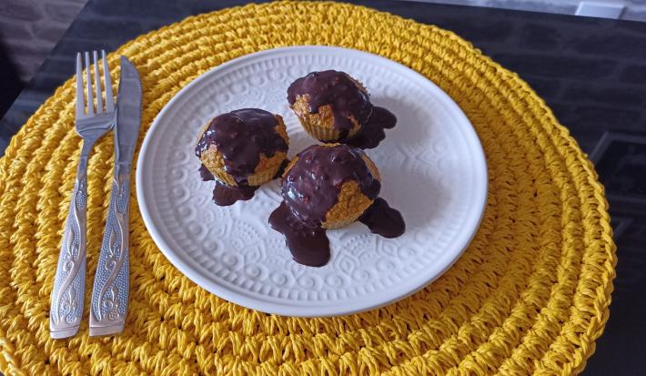 Fotografia em tons de amarelo em uma bancada de pedra, um porta prato amarelo de renda, um prato branco com bolinhos de cenoura com cobertura de chocolate por cima.