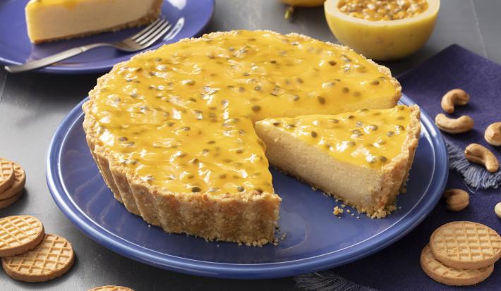 Fotografia em tons de cinza  e azul de uma bancada cinza com um prato redondo azul, sobre ela uma torta de maracujá. Ao fundo um prato azul com uma fatia de torta, um maracujá cortado ao meio. Ao lado biscoitos São Luiz e castanhas-de-caju espalhadas.