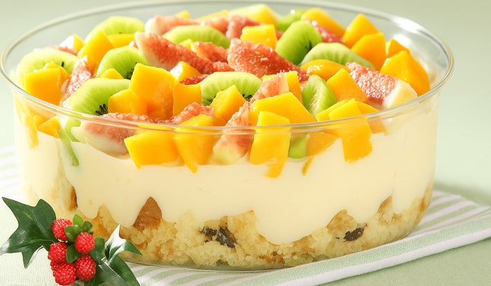 Fotografia em tons de amarelo, verde e vermelho em uma bancada de madeira verde clara, um pano branco com listras verdes, um recipiente de vidro com o pavê de panetone com frutas dentro dele.