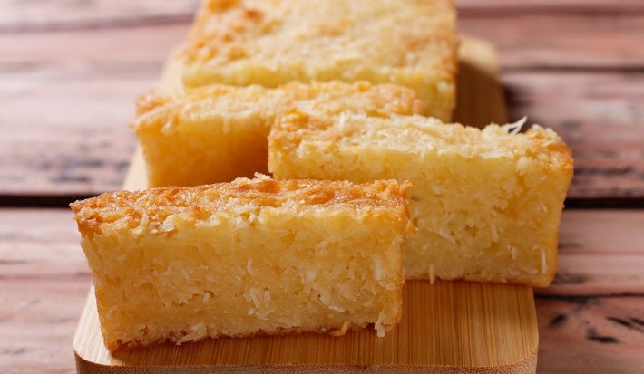 Bolo dourado de coco cortado em retângulos sobre tábua de madeira. Textura úmida e macia, com coco ralado por dentro e por cima. O fundo rústico de madeira realça o tom dourado da massa.
