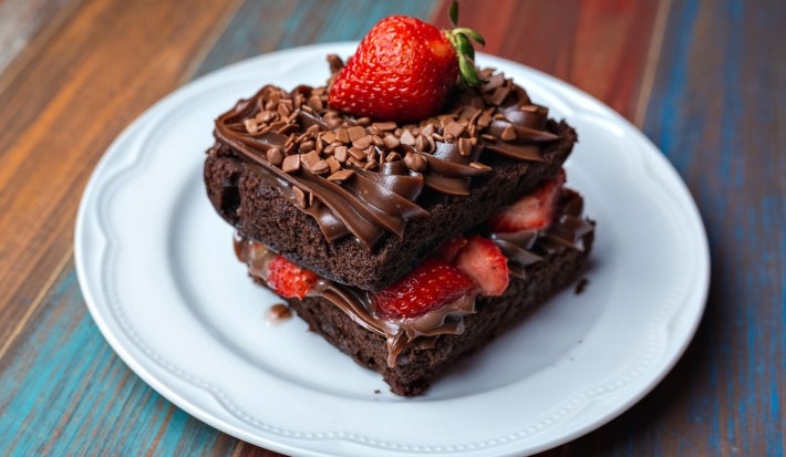 Sobre uma mesa, prato com uma fatia de bolo de chocolate. A massa é de chocolate, recheada com chocolates e morangos picados, coberta com chocolate e decorada com granulado e morangos cortados ao meio.