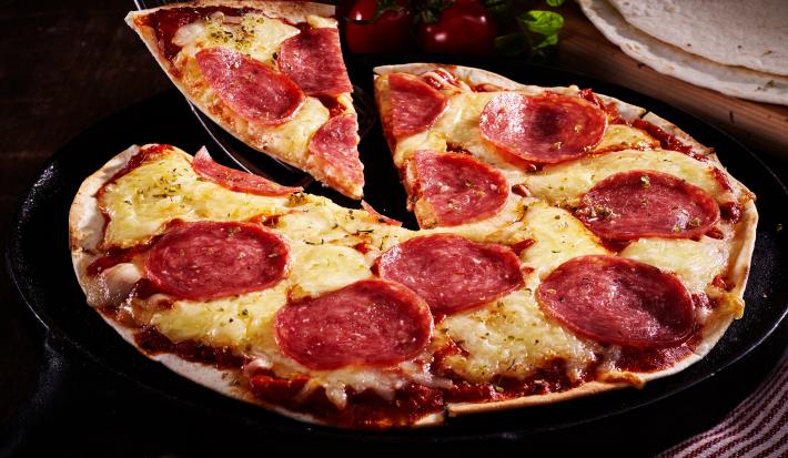 Fotografia em tons de marrom com uma pizza feita com massa de tortilha ao centro. A pizza é feita de queijo, salame e orégano.