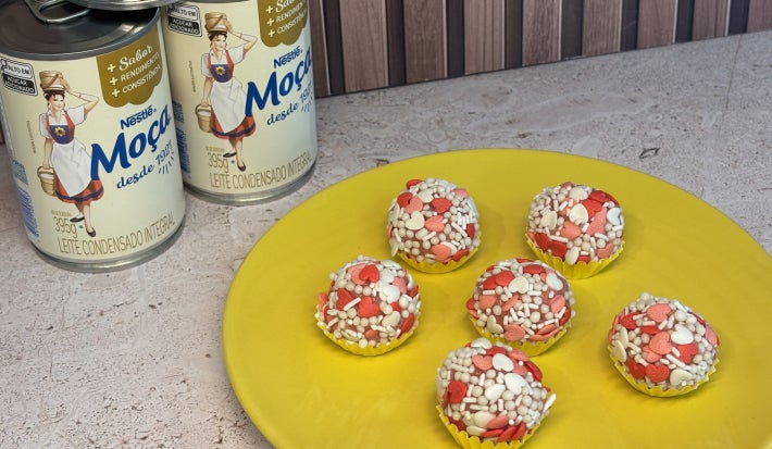 Docinhos decorados com confeitos brancos e vermelhos em formato de coração, sobre prato amarelo e com de Leite Moça ao fundo.