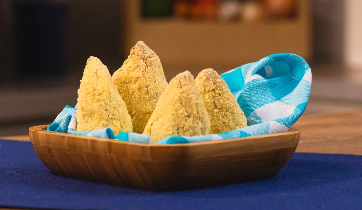 Fotografia de uma bancada de madeira com um pano azul escuro. Sobre ele tem um recipiente quadrado de madeira com um pano azul e branco forrando as quatro coxinhas saudáveis que estão dentro.