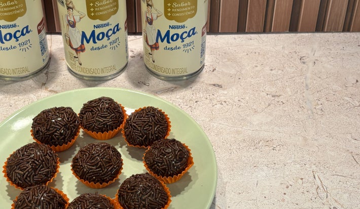 Brigadeiros cobertos com granulado de chocolate, em forminhas laranja sobre prato verde-claro, com Leite Moça ao fundo.