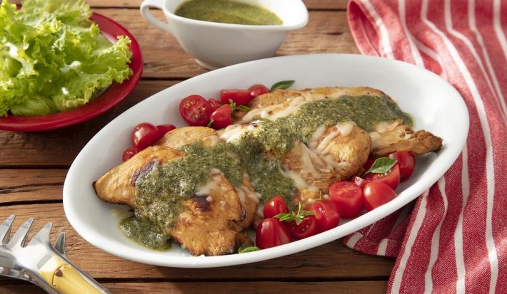 Foto da receita de frango caprese servida com tomates cereja em um prato largo e fundo de porcelana branca sobre uma mesa de madeira com um pano vermelho listrado a lado