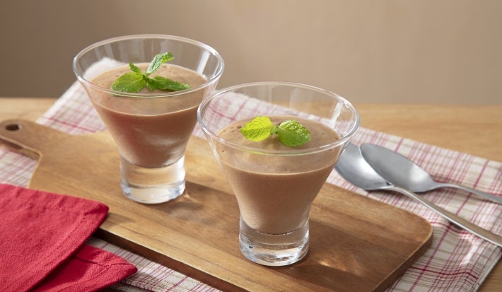 Fotografia de dois copinhos médios de vidro decorativos com a mousse sobre uma tábua de madeira retangular ao lado de duas colheres de sopa sobre um pano branco com listras vermelhas. Sobre as mousses tem folhas de hortelã.