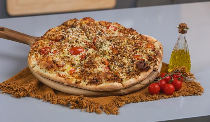 Foto da receita de massa básica de pizza, servida numa tábua de madeira com o sabor de pizza salgada. A tábua está ao lado de um galeteiro de azeite e alguns tomates.