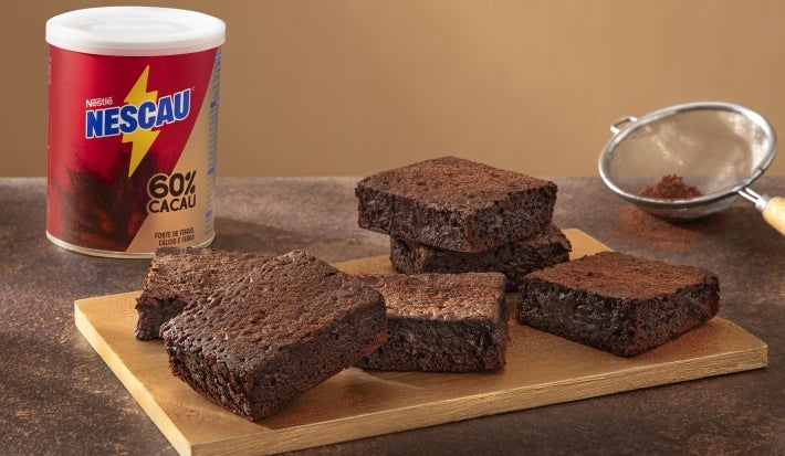 Foto da receita de brownie com nescau extra cacau servida em diversas porções sobre uma tábua de madeira, que acompanha uma peneira e uma embalagem de nescau ao fundo em cima de uma mesa de mármore escuro Foto da receita de brownie com nescau extra cacau servida em diversas porções sobre uma tábua de madeira, que acompanha uma peneira e uma embalagem de nescau ao fundo em cima de uma mesa de mármore escuro