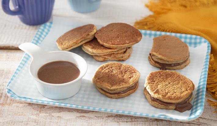 Foto em tons de marrom da receita de biscoito de panqueca servida em diversas porções sobre uma base quadriculada azul claro com um medidor com o recheio ao lado. Ao fundo há uma xícara azul e um pano laranja