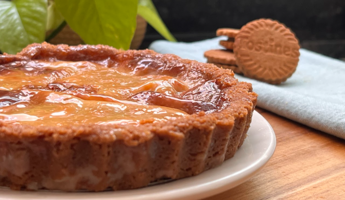 imagem mostra uma torta pequena sobre um prato branco. A torta em tons dourados com as bordas frisadas e um recheio caramelizado. Ao fundo um pano branco com 3 biscoitos Tostines e folhas verdes de uma planta.