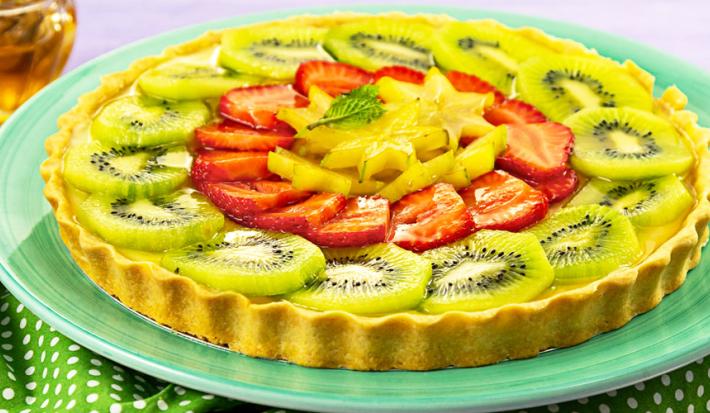 Fotografia em tons de verde e vermelho, com torta de frutas frescas fatiadas em prato verde sobre guardanapo verde com bolinhas brancas, potinho com mel ao fundo, tudo sobre bancada na cor cinza.