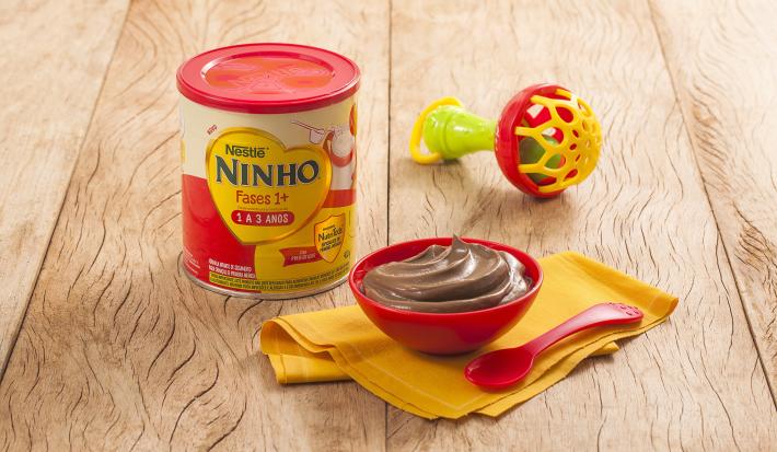 Fotografia em tons de amarelo e vermelho em uma bancada de madeira clara, um potinho vermelho em cima de um paninho amarelo, uma colher infantil vermelha e dentro do potinho um creminho de abacate com cacau. Ao fundo, brinquedinho e uma lata de Ninho Fase