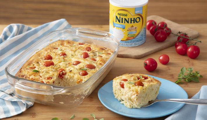 Fotografia em tons de azul de uma bancada de madeira com uma travessa de vidro com a torta cremoso, ao lado um pratinho azul com uma fatia de torta e ao fundo uma lata de NINHO Levinho e tomatinhos cereja.